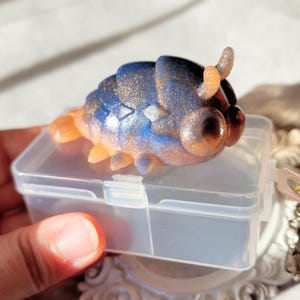 Op de afbeelding: Een klein, blauw en gouden harsfiguurtje van een beestje met grote ogen, dat in een doorzichtige plastic opbergdoos zit.