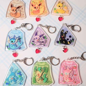 Puede incluir: Una colección de llaveros de acrílico transparente con varios personajes de dibujos animados. Cada llavero tiene un diseño único, con un dije en forma de corazón y un llavero plateado. Los personajes están en formas geométricas coloridas.
