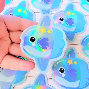 May include: A set of iridescent blue and purple holographic stickers featuring a cartoon moonfish with a tearful expression and yellow stars. The stickers are on a white wooden surface.