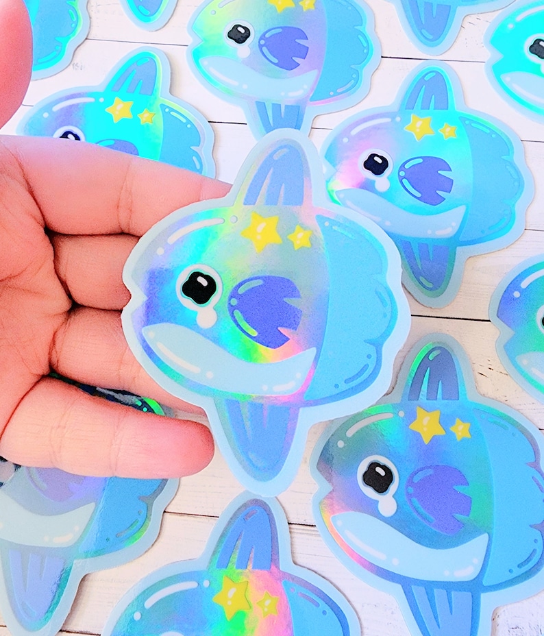 May include: A set of iridescent blue and purple holographic stickers featuring a cartoon moonfish with a tearful expression and yellow stars. The stickers are on a white wooden surface.