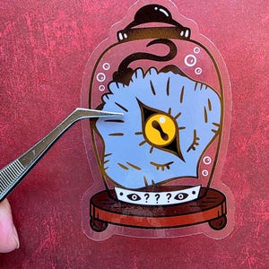 May include: A sticker featuring a blue, eye-shaped design inside a glass dome. The eye has a yellow iris and is surrounded by a blue, textured pattern. The base of the dome is brown, and the sticker is being held by tweezers.