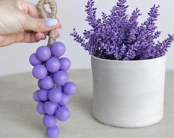 May include: A cluster of purple decorative grapes with a jute loop for hanging. Beside it, a white ceramic pot holds a vibrant arrangement of purple lavender. The items are set against a neutral background.