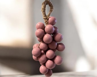 May include: A decorative cluster of pink or mauve spheres, resembling grapes, hangs from a woven loop. The spheres are tightly clustered, and the loop is made of a natural fiber. The background is blurred, highlighting the decorative object.