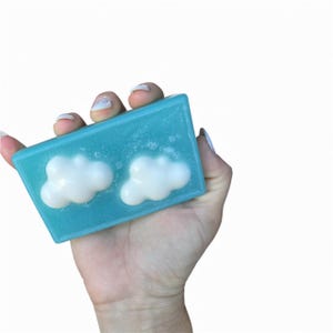 May include: A light blue rectangular soap bar with two white cloud-shaped decorations. The soap is held in a hand against a white backdrop, highlighting the design and colour of the soap.