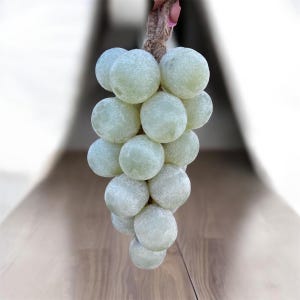 Puede incluir: Un racimo de uvas verde claro con aspecto esmerilado, sostenido por un tallo marrón. Las uvas son redondas y agrupadas, con una superficie ligeramente texturizada. El fondo es una superficie de madera borrosa.