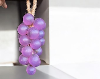 May include: A bunch of purple grapes made of a translucent material, hanging from a natural fiber rope. The grapes are a vibrant shade of purple, and the rope is a light brown color. The grapes are clustered together, creating a realistic appearance.