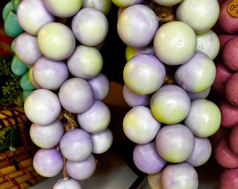May include: Two bunches of decorative grapes with a mix of green and purple spheres. The grapes are attached to a brown rope and are displayed on a black stand. The grapes have a smooth, painted finish.