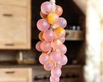 May include: A decorative hanging grape cluster made of translucent pink and amber-colored spheres. The spheres are strung together with a natural-colored twine, creating a unique and artistic home decor item. The background is blurred, suggesting an indoor setting.