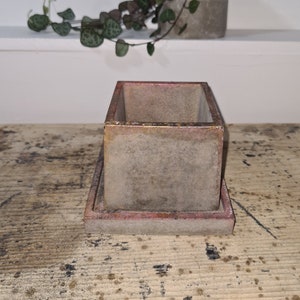 May include: A square concrete planter with a raised lip and a matching square base. The planter is light grey with a subtle pink and gold sheen.