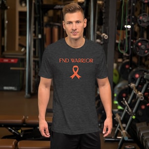 Puede incluir: Camiseta gris oscuro con el texto "FND WARRIOR" en naranja, junto con una cinta naranja. La camiseta de manga corta parece estar hecha de un material suave. La persona está en un gimnasio.