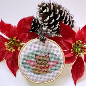 May include: A hand-stitched Christmas bauble featuring a brown cat with pink wings, surrounded by a green and white background. The bauble is framed in a wooden hoop and decorated with red poinsettia flowers.