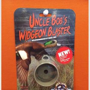 K&ouml;nnte beinhalten: Ein Jagdruf f&uuml;r Pfeifenten, mit der Aufschrift "Uncle Bob's Widgeon Blaster". Die Verpackung zeigt ein Foto einer fliegenden Pfeifente und den Text "NEW!" in einem roten Kreis. Der Ruf ist hellbraun.