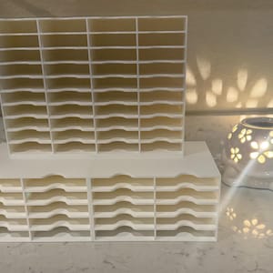 May include: A white plastic storage organizer featuring a tall vertical section with multiple rectangular slots and two shorter horizontal sections with stacked shelves, positioned next to a white ceramic turtle-shaped wax warmer.