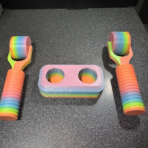May include: A set of 3D-printed items in a rainbow color scheme. The set includes two roller tools and a rectangular holder with two circular openings. The tools have a cylindrical roller and a handle. The holder is pink with rainbow edges.
