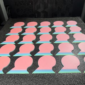 May include: A collection of pink and blue-green objects, possibly coasters or decorative pieces, arranged in rows on a dark surface. The objects have a rounded top and a layered, fan-like bottom. The color palette is soft and pastel.