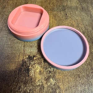 May include: A two-piece, round container with a lid. The lid and base are pink with a blue gradient on the sides. The interior of the lid is a square shape. The base is a light purple color.