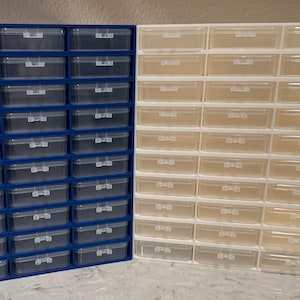 May include: Two rectangular storage organizers, one with a blue frame and one with a white frame, each featuring thirty individual clear plastic drawers arranged in a grid of three columns and ten rows, designed for sorting and storing small items.