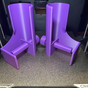 May include: Two matching purple 3D-printed objects. Each has a curved top, a horizontal threaded rod, and a knob. They are on a textured surface, likely a 3D printer bed. The objects appear to be designed for a specific purpose.