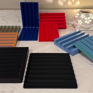 May include: A collection of colorful, grooved trays in various sizes and colors, including blue, red, black, orange, and green. The trays are arranged on a white countertop, showcasing their design and potential uses.