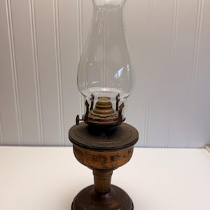 May include: An antique oil lamp with a clear glass chimney and a metal base. The lamp has a round base, a central stem, and a circular reservoir. The chimney is flared at the top and has a decorative edge. The lamp is made of metal and glass.