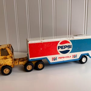 May include: Vintage Pepsi-Cola toy truck with a yellow cab and a trailer featuring the Pepsi logo in red, white, and blue. The truck shows signs of wear, adding to its nostalgic appeal. The trailer is white with red and blue accents.