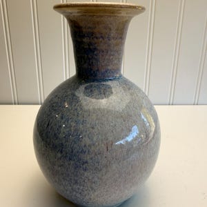 May include: A ceramic vase with a round body and a flared neck. The vase has a speckled blue and gray glaze with a brown rim. The vase is a decorative item.