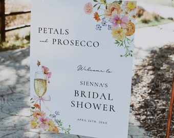 Puede incluir: Un cartel blanco con ilustraciones florales y el texto "Petals and Prosecco" en la parte superior. El cartel también dice "Welcome to Sienna's Bridal Shower" y "April 26th, 20XX". El cartel se muestra en un soporte de cobre.