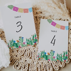 May include: Two white table number cards with a cactus and flower design. The top of each card has a string of colorful paper flags. The text "TABLE" is printed on each card, followed by the numbers "3" and "4".