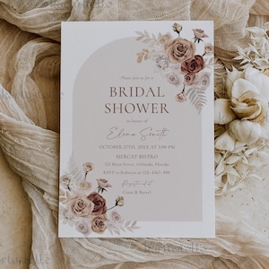 Puede incluir: Una invitación a una despedida de soltera con detalles florales en tonos marrón y crema. La invitación incluye el texto "BRIDAL SHOWER" y los detalles del evento. El diseño incorpora una forma de arco y está sobre un fondo de tela neutra.