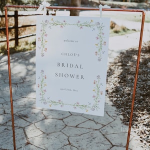 Może przedstawiać: Biały znak z kwiatową ramką i tekstem "welcome to CHLOE'S BRIDAL SHOWER april 26th, 20xx" jest wyświetlany na stojaku z miedzianej rury.
