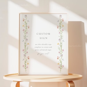 以下が含まれることがあります： 白い額縁に入ったポスターで、花柄の縁取りと黒い文字で「CUSTOM SIGN」と書かれています。下のテキストには「use this editable sign template to create and print unlimited signs for your event」と書いてあります。