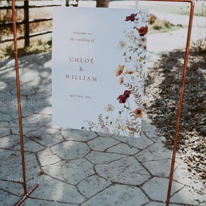 May include: A white wedding sign with a floral design in shades of red, orange, and white. The sign reads "Welcome to the wedding of Chloe & William, April 26th, 20xx". The sign is hanging from a copper pipe stand.