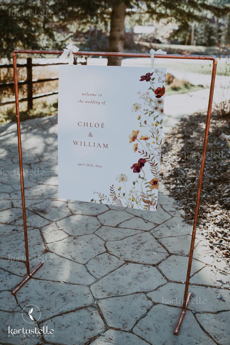 May include: A white wedding sign with a floral design in shades of red, orange, and white. The sign reads "Welcome to the wedding of Chloe & William, April 26th, 20xx". The sign is hanging from a copper pipe stand.
