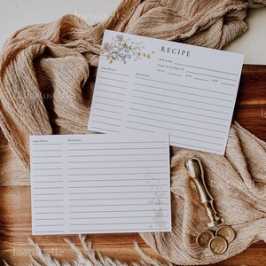May include: Two white recipe cards with floral accents and lined spaces for ingredients and directions rest on a wooden tray. A gold-colored wax seal stamp and three gold coins are also present, along with a beige fabric.