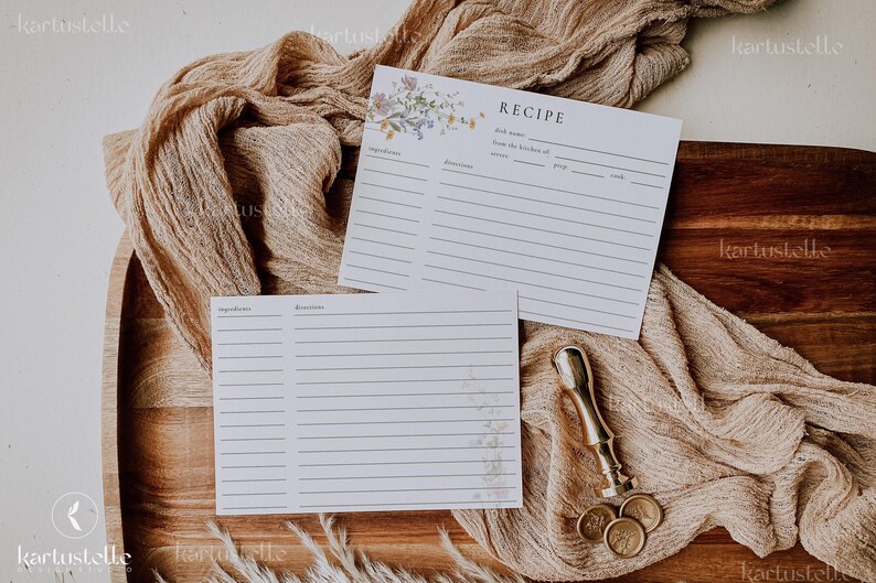 May include: Two white recipe cards with floral accents and lined spaces for ingredients and directions rest on a wooden tray. A gold-colored wax seal stamp and three gold coins are also present, along with a beige fabric.