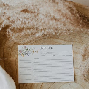 May include: A white recipe card with floral accents and the word "RECIPE" at the top. The card includes spaces for ingredients, directions, and other details. The background features dried plants and a wooden surface.
