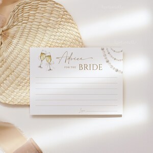 May include: A white card with gold lettering that says "Advice for the Bride". There are two champagne glasses with gold rims and a string of pearls on the card. The card is on a white background with a woven straw fan and a beige ribbon.
