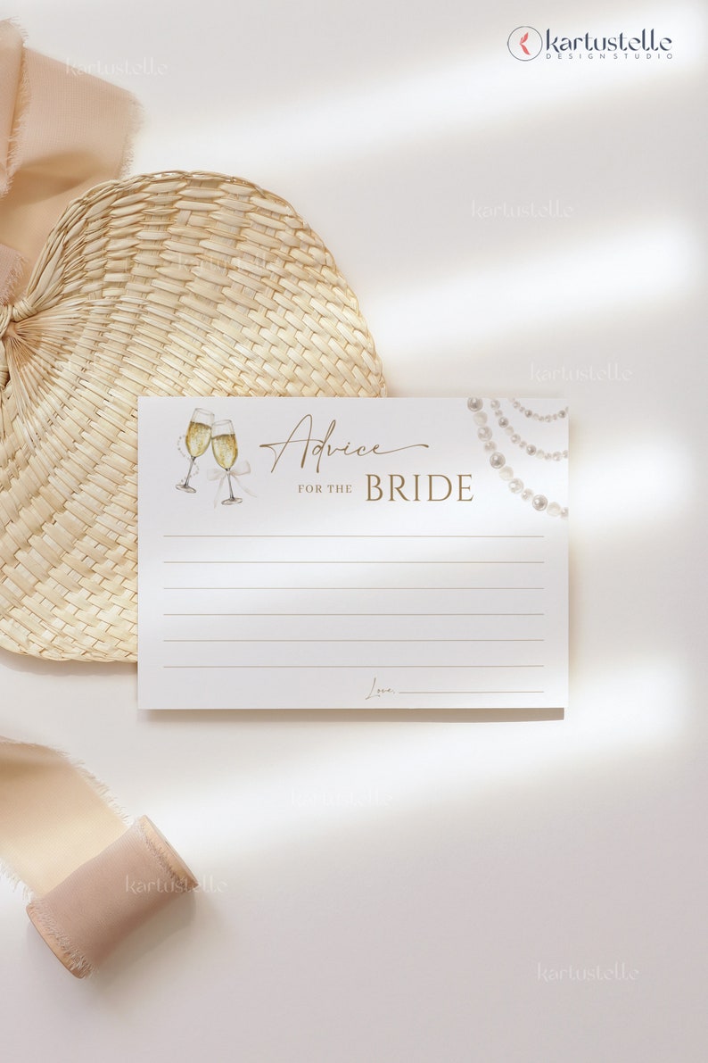 May include: A white card with gold lettering that says "Advice for the Bride". There are two champagne glasses with gold rims and a string of pearls on the card. The card is on a white background with a woven straw fan and a beige ribbon.