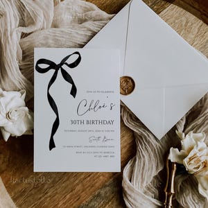 K&ouml;nnte beinhalten: Eine wei&szlig;e Einladungskarte mit schwarzem Schleifendesign und dem Text "Chloe's 30th Birthday". Ein Umschlag mit goldenem Siegel und ein Holztablett mit Stoff und Blumen vervollst&auml;ndigen das elegante Design.
