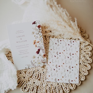 May include: A wedding invitation with a floral design featuring burgundy, orange, and white flowers. The invitation is for Chloe and William's wedding on October 27th, 20XX. The invitation is on a beige crocheted placemat with a white fluffy feather plume.