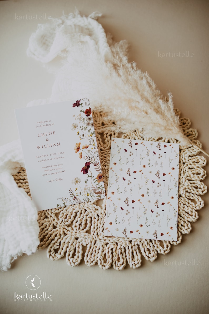 May include: A wedding invitation with a floral design featuring burgundy, orange, and white flowers. The invitation is for Chloe and William's wedding on October 27th, 20XX. The invitation is on a beige crocheted placemat with a white fluffy feather plume.