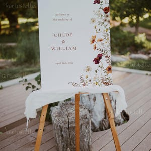 May include: A white wedding welcome sign with a floral design featuring burgundy, orange, and yellow flowers. The sign reads "Welcome to the wedding of Chloe & William, April 26th, 20xx".