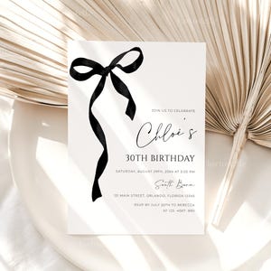 K&ouml;nnte beinhalten: Eine wei&szlig;e Einladungskarte mit einer schwarzen Schleifenillustration und Text. Die Einladung lautet "Chloe's 30. Geburtstag" mit Veranstaltungsdetails. Die Karte liegt vor einem neutralen Hintergrund mit einem getrockneten Palmblatt.