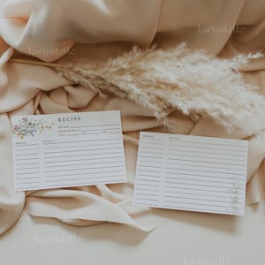 May include: Two white recipe cards with lined spaces for ingredients and directions. One card has floral accents and the word "RECIPE" at the top. The cards rest on a beige fabric with a dried plant stem.