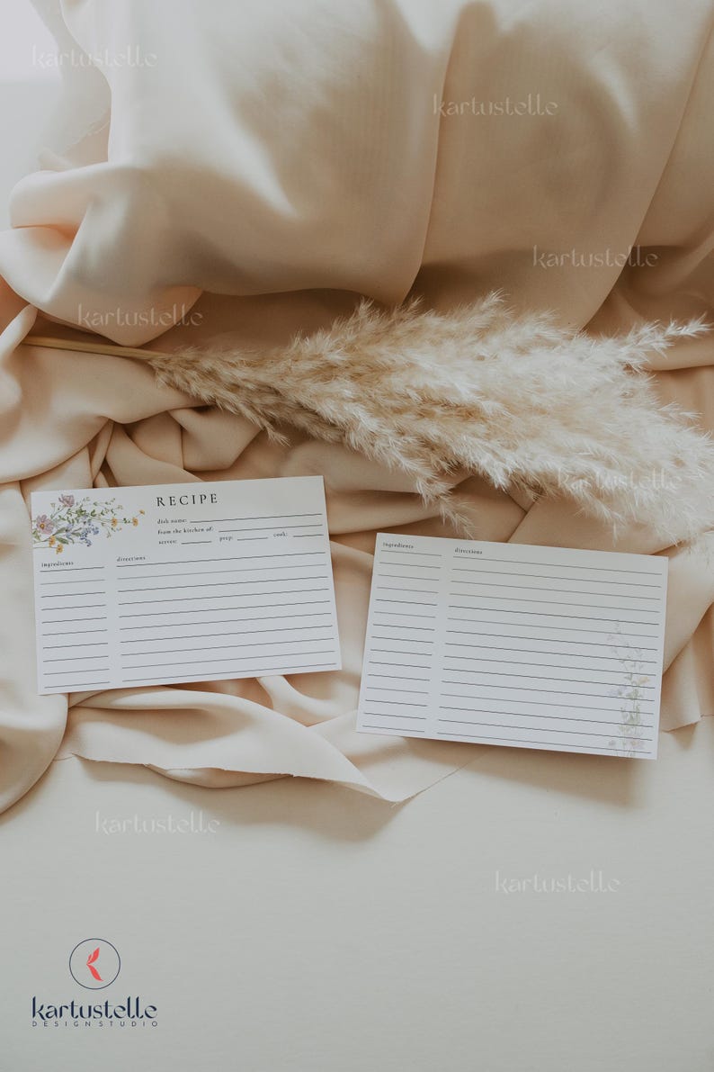 May include: Two white recipe cards with lined spaces for ingredients and directions. One card has floral accents and the word "RECIPE" at the top. The cards rest on a beige fabric with a dried plant stem.