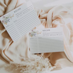 May include: Two white recipe cards with floral accents and lined spaces for ingredients and instructions. The word "RECIPE" is at the top of each card. The cards are on a beige fabric background.