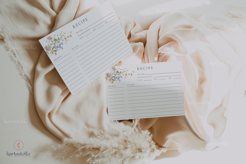 May include: Two white recipe cards with floral accents and lined spaces for ingredients and instructions. The word "RECIPE" is at the top of each card. The cards are on a beige fabric background.