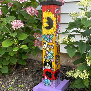 May include: A colorful wooden garden stake with a black cat, sunflowers, and flowers painted on it. The stake has a purple base and a solar-powered light on top. The text "MEOW" is painted on the stake.