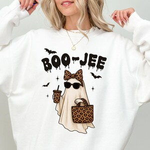 Op de afbeelding: Witte sweatshirt met een Halloween-thema. Het ontwerp toont een spook met een zonnebril en een luipaardprint strik, met een koffiebeker en een handtas. De tekst "BOO-JEE" staat boven het spook.