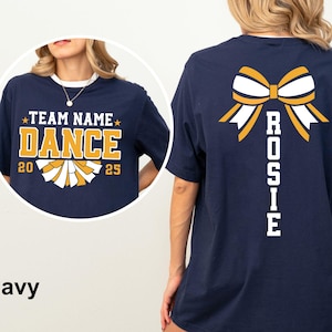 May include: Navy blue t-shirt with a cheerleading design. The front features the text "TEAM NAME DANCE 20 25" in gold and white. The back displays a gold and white bow graphic with the name "ROSIE" in white.